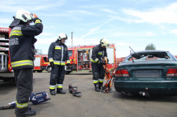 Három tűzoltó erős támaszt tesz egy autó oldalához háttérben tűzoltóautó