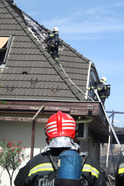 Háttal egy tűzoltó piros sisakban előtte a háttérben tetőn dolgoznak a többiek