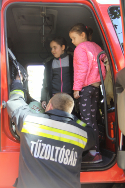 Tűzoltóautóban két kisgyerek áll előttük háttal egy tűzoltó beszél hozzájuk