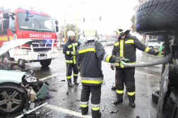 Balra tűzoltóautó előtte autó összetört eleje jobbra tűzoltók kötelet húznak ki a felborutl autó alvázától
