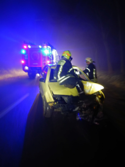 Összetört autó szemből a motorháztetőn egy tűzoltó mögötte a társa és egy tűzoltóautó a háttérben