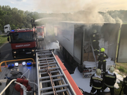 Kigyulladt kamion mellett tűzoltóautó tetején tűzoltó áll és vízsugárral olt miközben a többi tűzoltó a kamion hátuljánál dolgozik